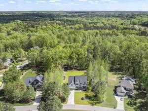 Aerial view of a suburban neighborhood with several houses, green lawns, and driveways, surrounded by dense forest and trees under a clear blue sky.