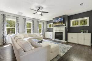 A bright living room with a large sectional sofa, a patterned rug, a fireplace with a TV above it, built-in cabinets, and multiple windows with patterned curtains letting in natural light.