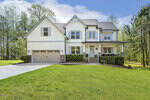 Charming two-story white house at 1723 River Clb Way, Franklinton features a gray roof, double garage, covered front porch with chairs, American flag, and a spacious front lawn; nestled among trees under a blue sky with clouds.