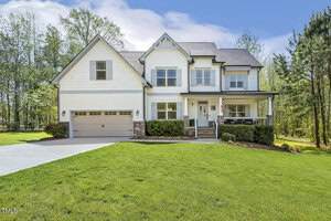 Charming two-story white house at 1723 River Clb Way, Franklinton features a gray roof, double garage, covered front porch with chairs, American flag, and a spacious front lawn; nestled among trees under a blue sky with clouds.