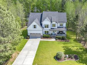 A two-story light blue house with dark shingles, a covered front porch, and an attached two-car garage, sits on a large green lawn with surrounding trees and a concrete driveway.