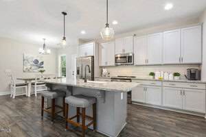 A modern kitchen with white cabinets, stainless steel appliances, a gray island with a granite countertop, pendant lights, and two stools. A dining table with chairs is visible in the background near a window.