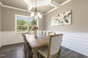 A dining room with a wooden table, six chairs, a vase with pink flowers, a large window showing greenery outside, a floral painting on the wall, and a modern light fixture hanging from a coffered ceiling.