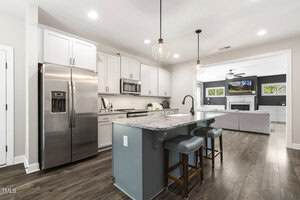 Modern kitchen with white cabinets, stainless steel appliances, granite island with sink, two stools, pendant lights, and a view into a living room with a fireplace and TV. Hardwood floors throughout the space.
