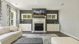 A living room with a white sectional sofa, a gray ottoman, a blue patterned rug, and a fireplace centered between built-in cabinets. A landscape painting hangs above the mantel; large windows let in natural light.