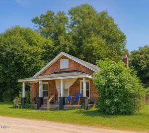A small brick house with a covered front porch featuring two blue chairs, a table, and curtains. There is a grassy yard, a dirt road in front, and large leafy trees in the background.