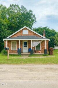 A small brick house with a covered front porch, three blue chairs, a wooden rocking chair, a lace curtain, a red mailbox, and a brown trash bin in front, surrounded by green grass and trees.