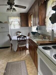A kitchen with wood cabinets, white appliances, a table covered with a white cloth and four chairs. Light enters through small windows with lace curtains. Dishes are drying on a rack next to the sink.