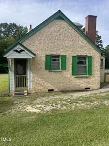 A small beige stucco house with a steep roof, green shutters, a brick chimney, two front windows, and a screened front door. The house sits on a grassy lawn with some patchy areas and trees in the background.