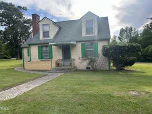 A small, beige house with a sloped green roof, green window shutters, and a front porch with two chairs. The house sits on a grassy lawn with a curved sidewalk and a large bush near the entrance.