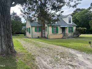 A beige house with green shutters and a dormer window sits next to a gravel driveway at 45 Perkinson Street in Kittrell, surrounded by grass and trees under a mostly cloudy sky.