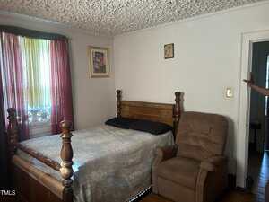 A bedroom with a wooden bed, two black pillows, a light-patterned bedspread, a brown recliner chair, a window with curtains, and two pictures on the wall. A person's arm is visible near the doorway.