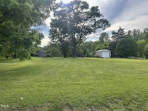 A large grassy yard with neatly mowed lines, bordered by trees, with a white shed and a smaller gray shed in the background under a partly cloudy sky.