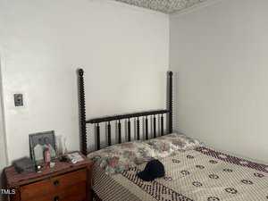 A bedroom with a wooden four-poster bed, a floral and checkered quilt, and a nightstand. The nightstand holds a framed photo, a lamp, a water bottle, lotion, and a black cap is on the bed.