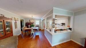 A dining area with a rectangular table and chairs is adjacent to a kitchen with white cabinets and countertops, separated by a half-wall. The space has wood floors and light-colored walls with decorative items on display.