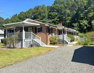 A one-story brick house with a covered front porch, white railings, and black shutters sits beside a gravel driveway. The yard has bushes, lawn, and is bordered by tall trees under a clear blue sky.