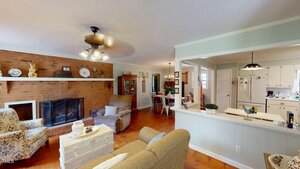 A cozy living room with a brick fireplace, armchairs, and a chest-style coffee table; an open layout leads to a dining area and a white kitchen, all with wood flooring and light-colored walls.
