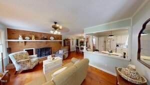 A living room with wood floors, a brick fireplace, armchairs, a sofa, and decorative items. An open window-style cutout in the wall leads to a white kitchen. Ceiling fan with lights is on the ceiling.