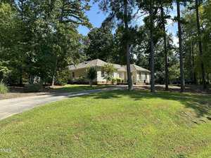 A single-story yellow house with white trim sits among tall trees. The house has a gray roof and a curved driveway, with green grass and landscaped plants in the front yard. The weather appears sunny.