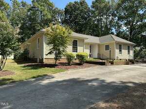 Charming single-story yellow house with white trim at 3002 Sidney Hill in Henderson. Features a gray shingle roof, multiple windows, concrete driveway, landscaped bushes, and trees beneath a clear blue sky.