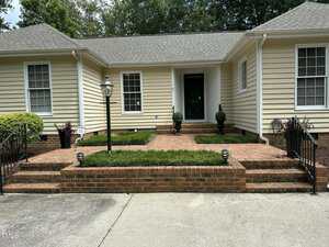 Single-story beige house with white trim, a brick porch and steps, black metal railings, and a black lamp post. Two potted plants flank the front door. Trimmed shrubs line the walkway and porch area.