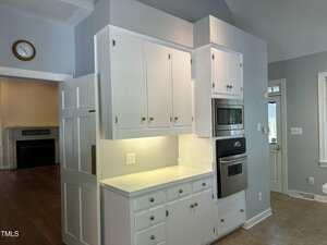 A kitchen area with white cabinets, a built-in microwave and oven, a countertop with under-cabinet lighting, and a white door. An adjacent room with a fireplace and a wall clock is partially visible.