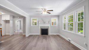 A bright living room with light gray walls, a ceiling fan with lights, wood-style flooring, a decorative fireplace, and three large windows letting in natural light. Doorways lead to other rooms and a kitchen area.
