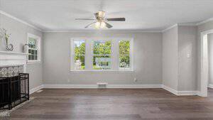 A bright living room with light gray walls, three large windows, a ceiling fan with lights, a fireplace with a decorative screen, and wood-look flooring. White trim outlines the windows, doors, and ceiling.