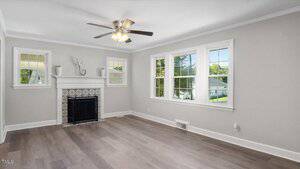 A bright living room with light gray walls, wood flooring, a ceiling fan with lights, a fireplace with a white mantel and decorative tile, and three large windows letting in natural light.