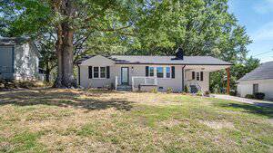 This charming single-story house at 304 Lanier Street in Oxford features light-colored siding, a small front porch, black shutters, and a convenient carport on the right. Large trees provide shade over the straw-covered lawn.