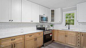 A modern kitchen features white upper cabinets, light wood lower cabinets, a stainless steel microwave, and oven, white subway tile backsplash, a black faucet, and a window above the sink.