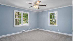 A room with light blue walls, two white-trimmed windows, light gray carpet, and a ceiling fan with lights. The room is empty and sunlight comes in through the windows.