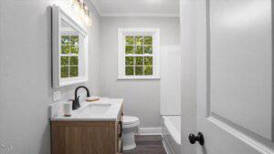 A small bathroom with a white door, a wooden vanity with a white countertop and black faucet, a mounted mirror, a toilet, a bathtub with shower, and a window letting in natural light.