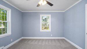 Empty room with light blue walls, gray carpet, white trim, a ceiling fan with lights, and two windows letting in natural light. The room appears clean and freshly finished.