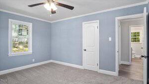 A small empty bedroom with light blue walls, white trim, a window, two white doors, beige carpet, a ceiling fan with lights, and an adjoining bathroom visible through an open door.