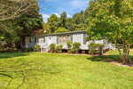 A single-story, light gray manufactured home with white trim sits on a well-maintained lawn at 1476 Tollie Weldon Road in Henderson. Bushes line the front, a small porch with steps welcomes you, and trees surround the property.