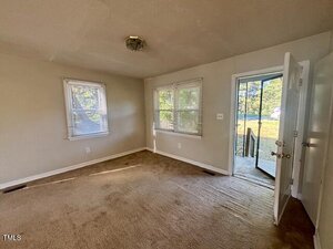 Empty room with beige carpet, light gray walls, two windows, and an open white door leading outside to a small porch with steps. Sunlight filters through the windows, and a ceiling light fixture is visible.