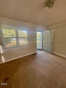 A small room with beige carpet and light-colored walls, featuring a window with blinds on the left and a front door partially open to the outside. Sunlight is coming through the window.