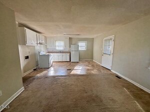 An unfurnished room with beige carpet leading into a small kitchen area with white cabinets, a white stove, and a white refrigerator. Two windows and a back door are visible, and the walls are painted light beige.