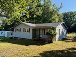 Charming single-story gray house at 1708 Monarch Drive in Creedmoor, featuring a covered front porch with red shutters and a small bush by the steps. A white trailer sits in the background amid lush grass and trees.