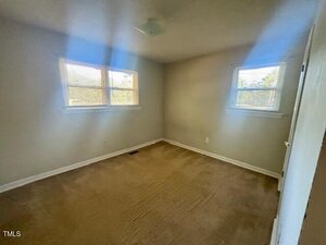 A small, empty bedroom with beige carpet, off-white walls, and two windows with blinds letting in natural light. There is a ceiling light fixture and a closed white door is visible on the right.