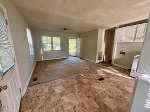 A small, unfurnished living space with a mix of carpet and vinyl flooring, white walls, and windows on two sides. A door leads outside, and the kitchen area is partially visible with upper cabinets and appliances.