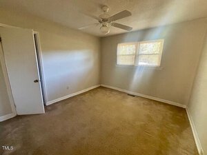 Empty room with beige carpet, off-white walls, a white ceiling fan, a partially open white door, and two windows with blinds letting in natural light from the right wall.