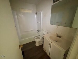 Small bathroom with a bathtub and shower combo, white toilet, white vanity with sink, wall-mounted mirrored cabinet, and wood-look flooring. The space appears compact and minimally decorated.