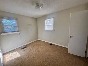 Small, empty room with beige carpet, light-colored walls, two windows with blinds, and an open door. Electrical cords are on the floor near one wall, and sunlight comes in through the left window.