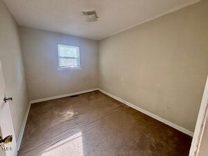 Small empty room with beige walls, a brown carpet, a single window with blinds, and a ceiling light fixture. Sunlight enters through the window, casting shadows on the floor.