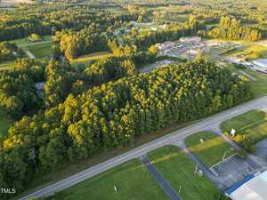Aerial view of a tree-covered area bordered by a paved road in the foreground, with grassy fields, scattered buildings, and a residential development in the background. Dense forest occupies most of the center of the image.