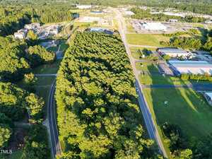 Aerial view of a forested area bordered by roads near 01 158 Business Hwy, Henderson, with commercial and industrial buildings, parking lots, and open grassy fields surrounding the woods. The landscape stretches to the horizon under bright daylight.