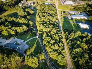 Aerial view of a winding road cutting through a dense, green forest area, with buildings, parking lots, and grassy fields visible on either side. Sunlight casts shadows from the trees across the landscape.