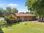Charming single-story brick house at 4642 Fielding Knott Road, Oxford, with a brown roof, white front door, and several windows. The yard features green grass, a few trees, and a wooden ramp to the side entrance under blue skies.